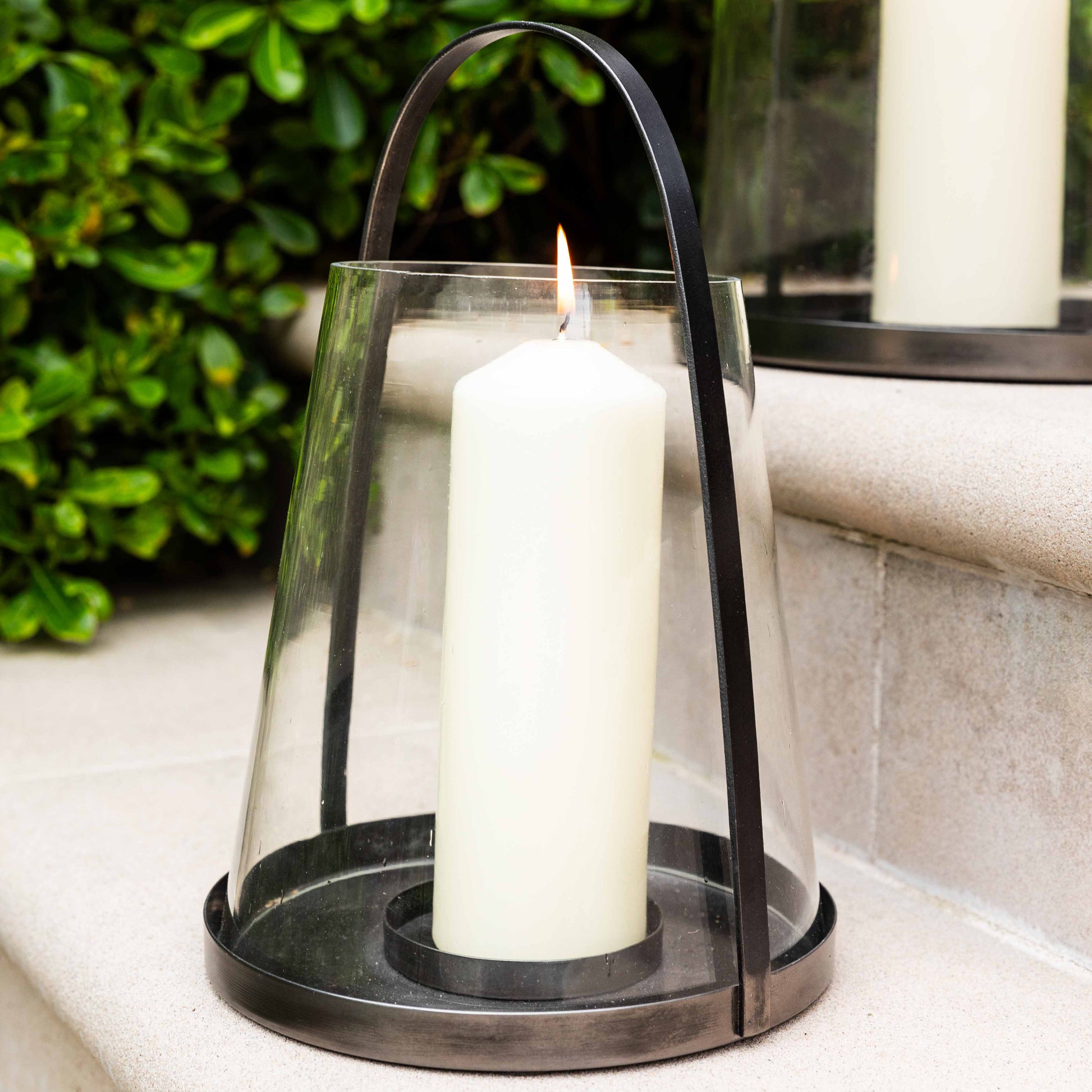 Candle in a glass lantern with a black metal handle against a green leafy background