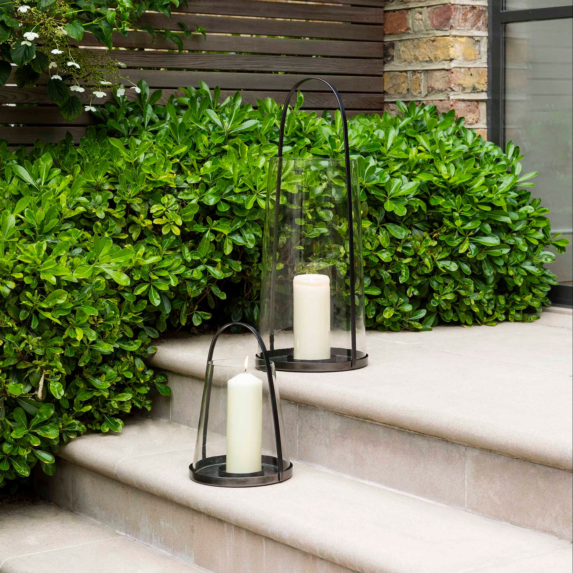 Candle holders lanterns modern black coated metal with handle and glass dome surround with candles on outdoor steps surrounded by greenery