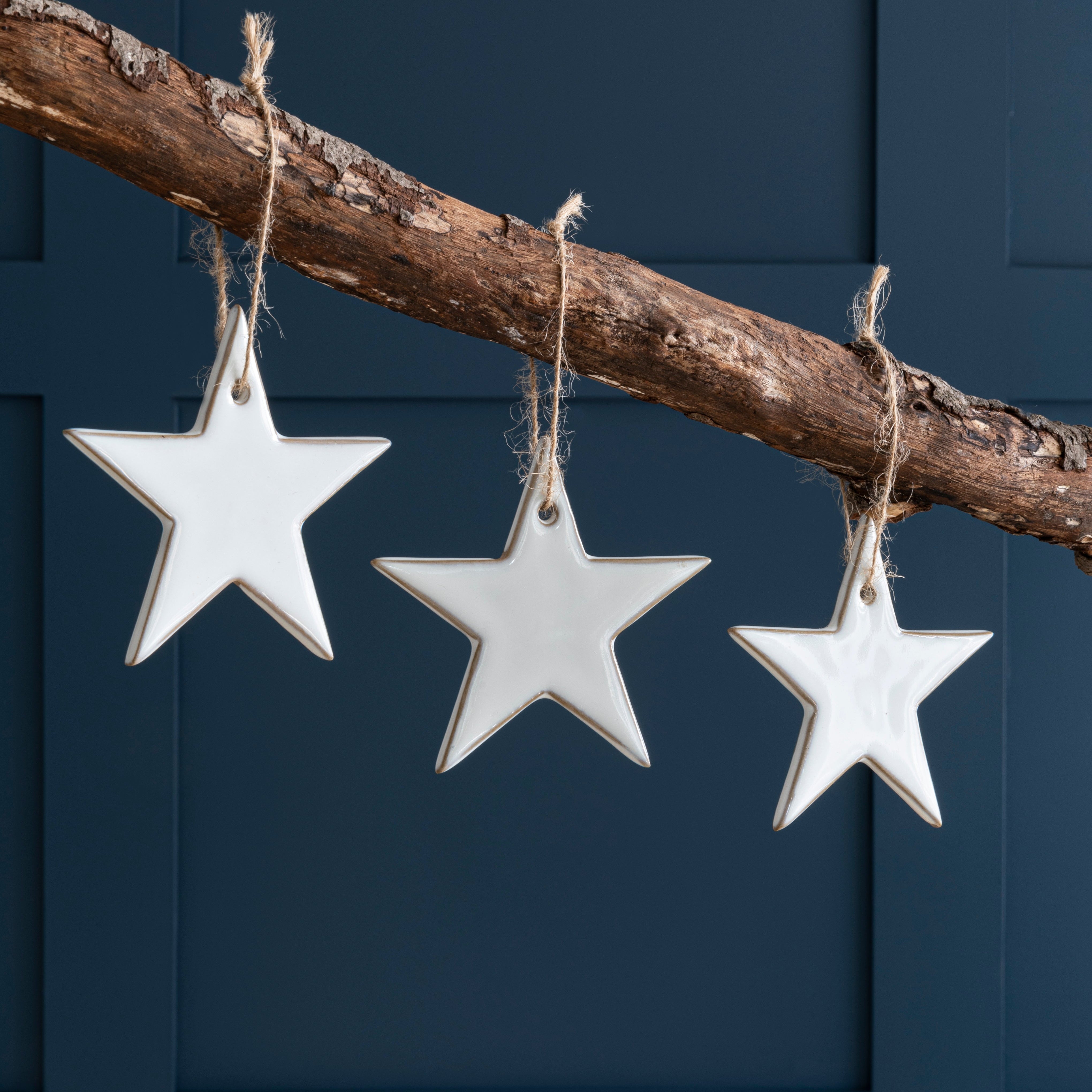 A set of three white ceramic stars with a glaze finish, hanging from a jute loop against a dark blue background.