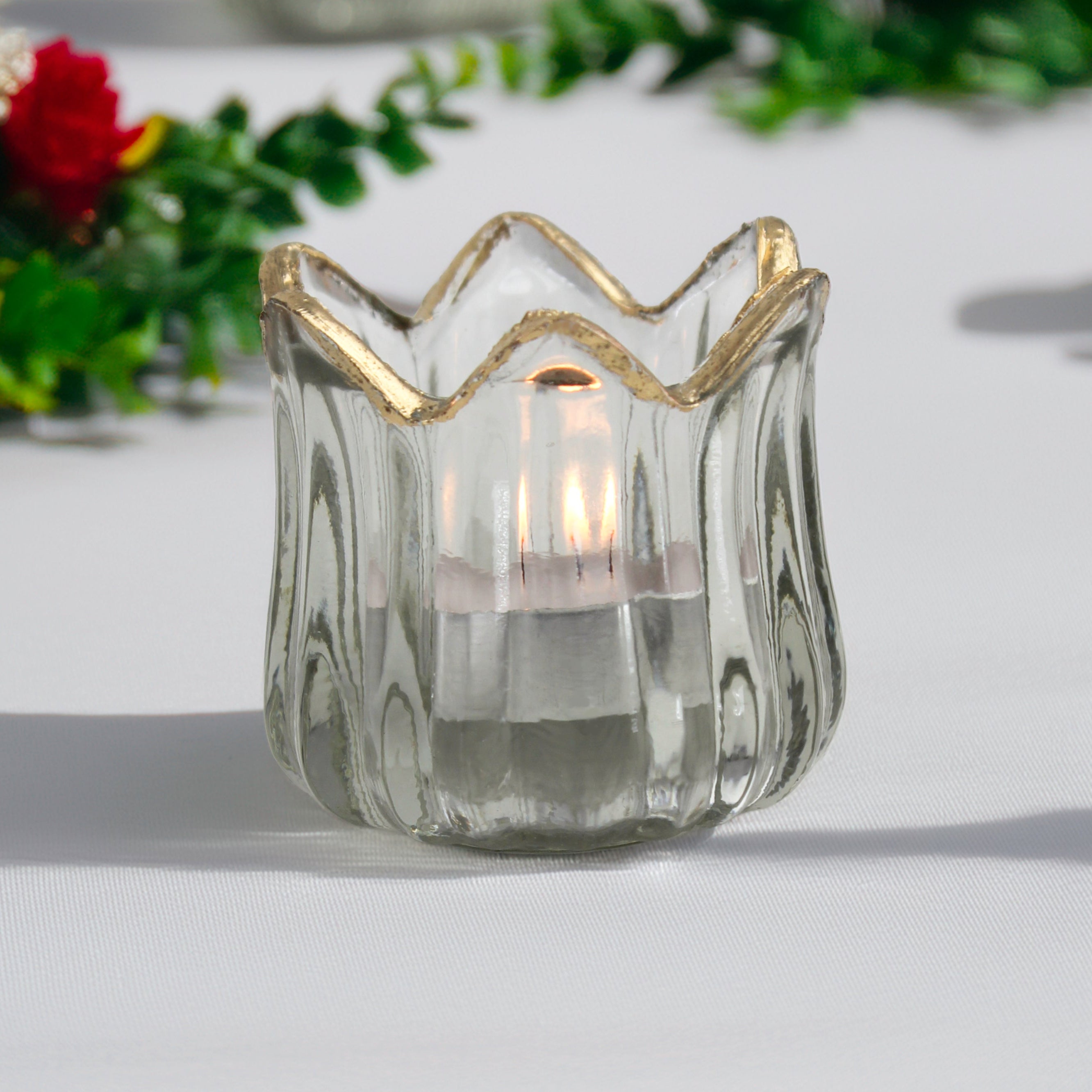 Clear glass candle holder with gold rim on a white surface with blurred flowers in the background