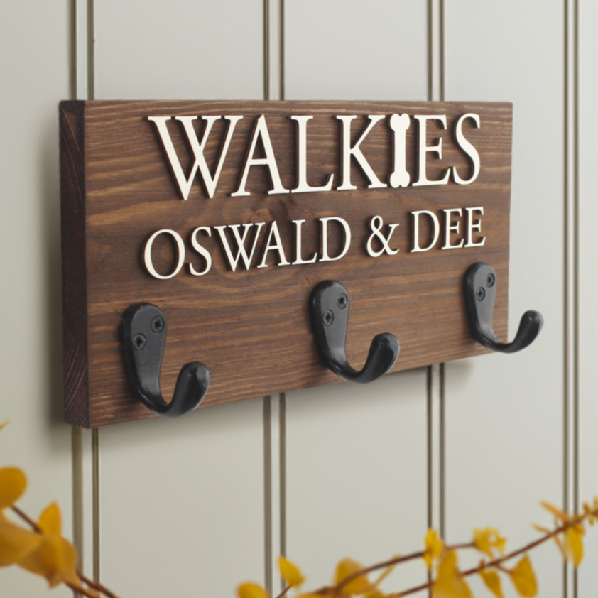 Our personalised dog lead hook with three sturdy hooks is handcrafted from premium wood, offering a stylish and functional storage solution for your pet's leads, collars, and accessories. Available in eight stunning wood finishes, it features raised lettering in natural wood or black, with the option to add your pet’s name and a paw print for a personal touch. Perfect for organising your pet’s gear in style. this image is dark walnut with raised natural wood wording with paws