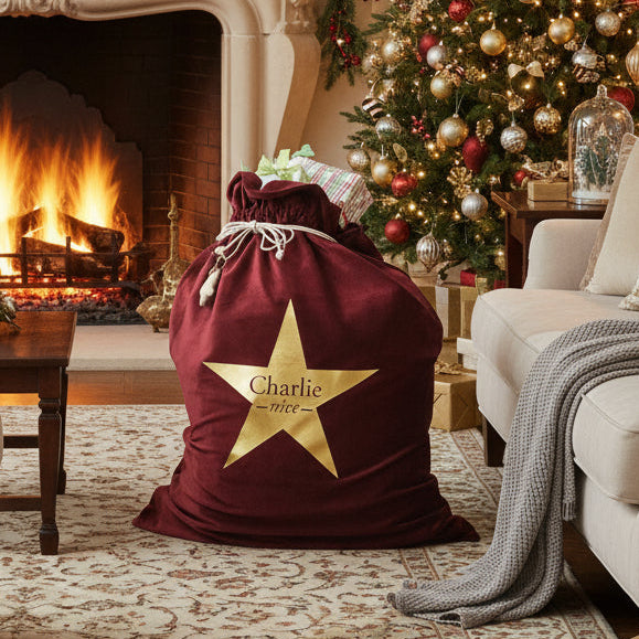A red velvet Santa sack with a golden star design and a personalized name 'Charlie' in gold lettering at the bottom. The sack is tied at the top with a white ribbon.