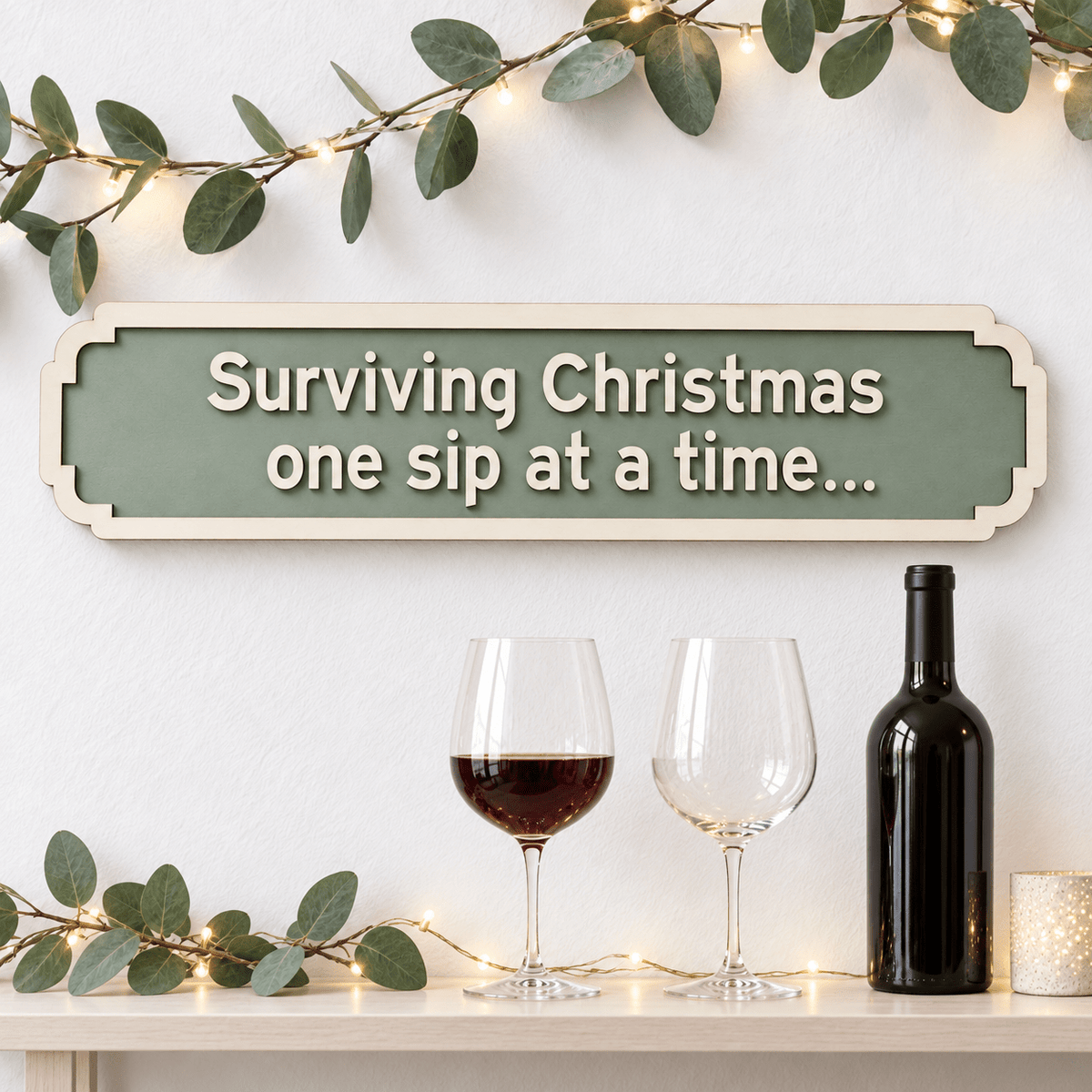 Sage green Christmas sign with raised wooden lettering reading “Surviving Christmas one sip at a time”, styled above wine glasses with festive greenery.