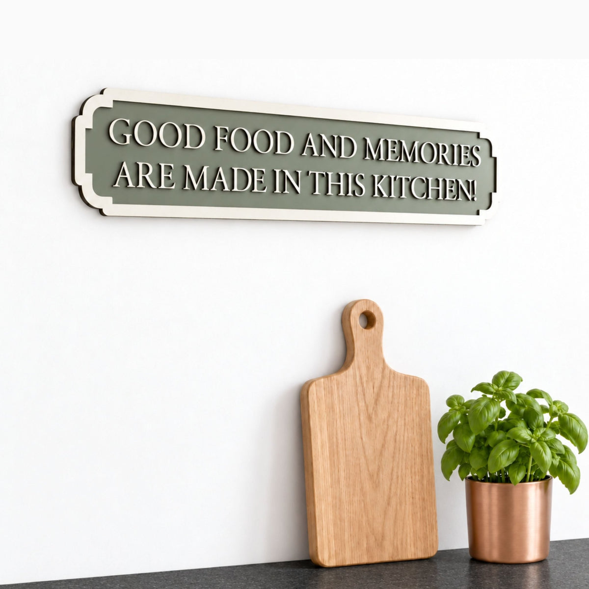 Green wooden kitchen sign reading “Good food and memories are made in this kitchen”, styled above chopping boards and a potted herb.