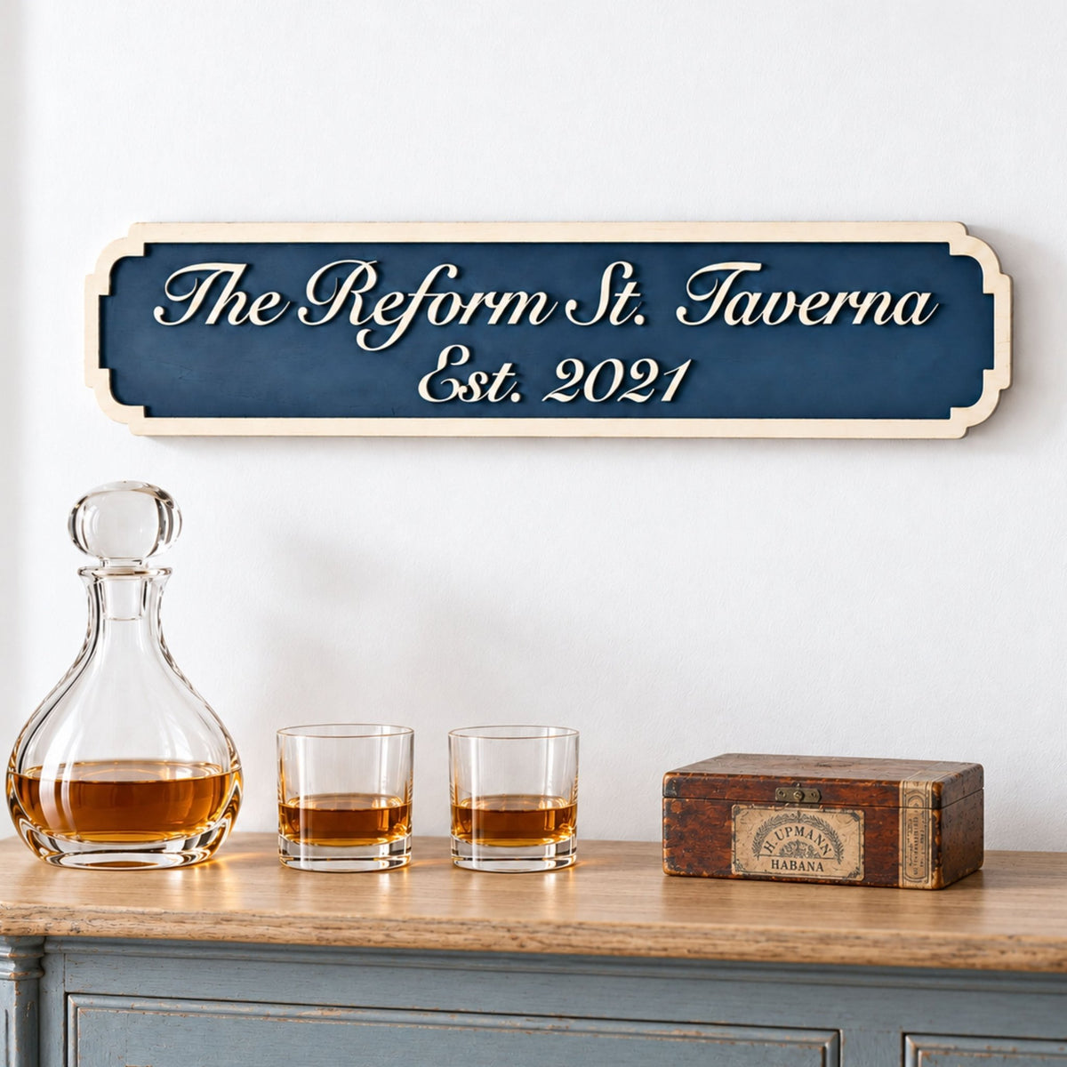 Blue personalised tavern sign with raised wooden script and an established date, styled above a drinks cabinet with a decanter and glasses.
