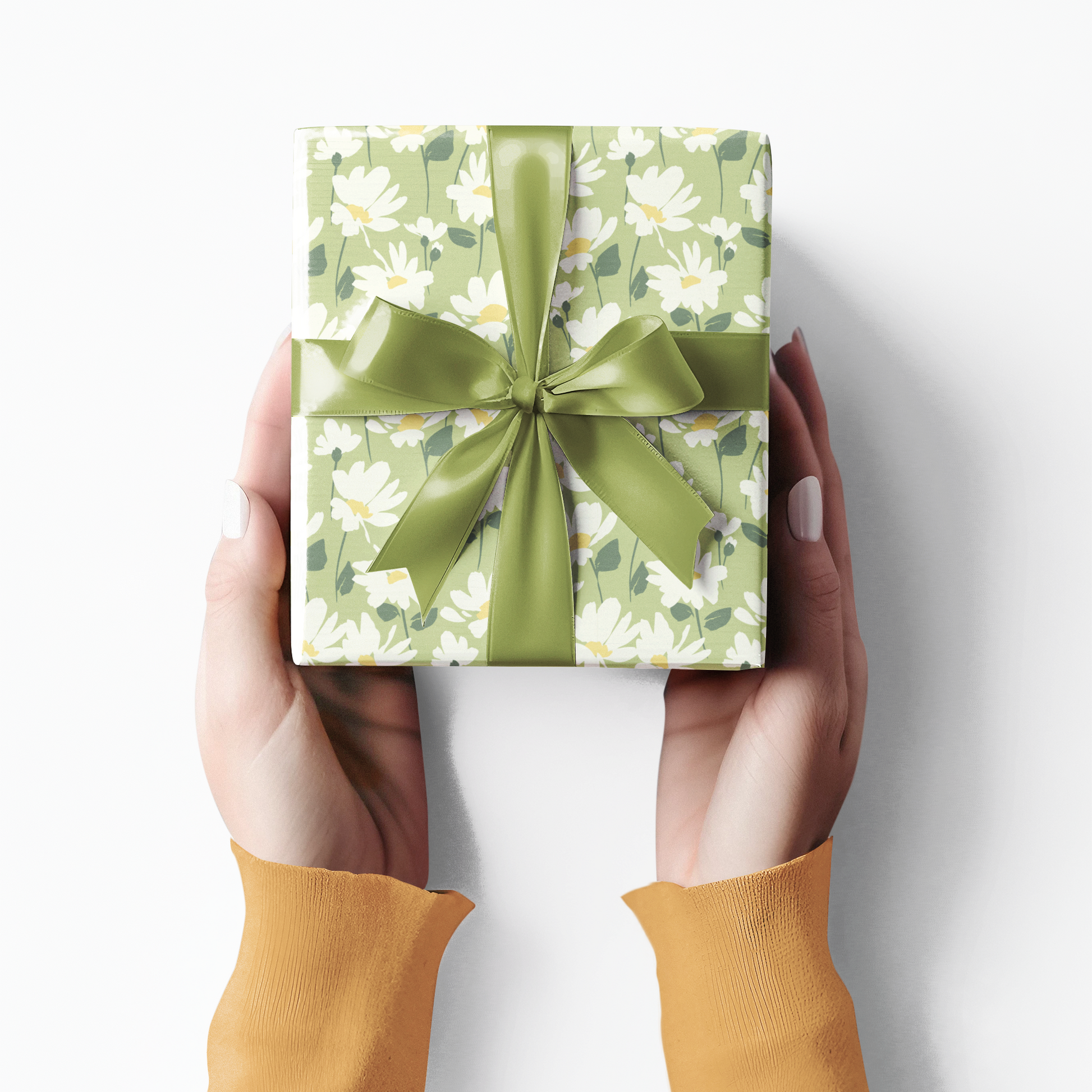 Gift box with green floral pattern and bow held by hands wearing mustard yellow sleeves on a white background