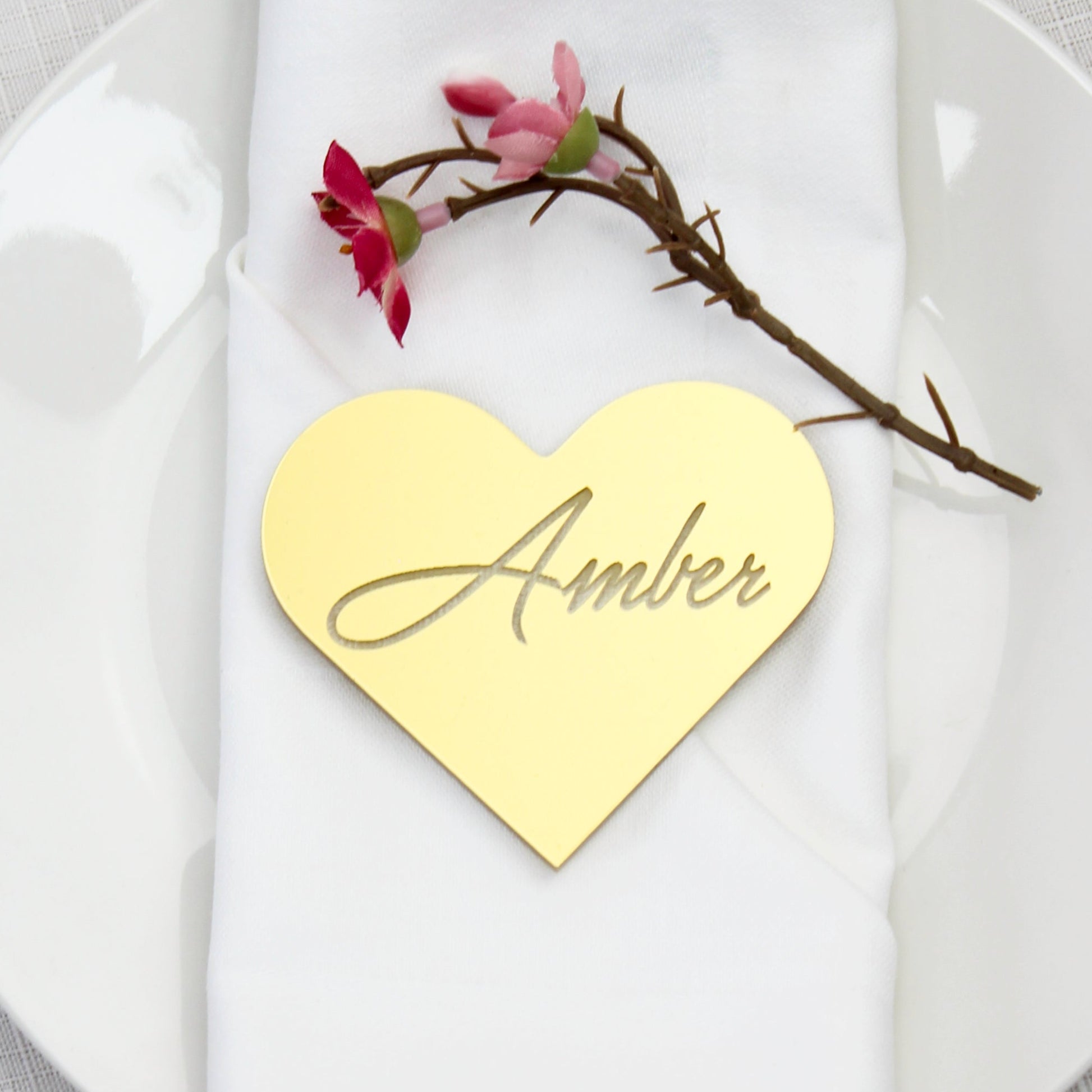 Heart-shaped place card with 'Amber' on a white napkin with a branch and flowers on a gray tablecloth.