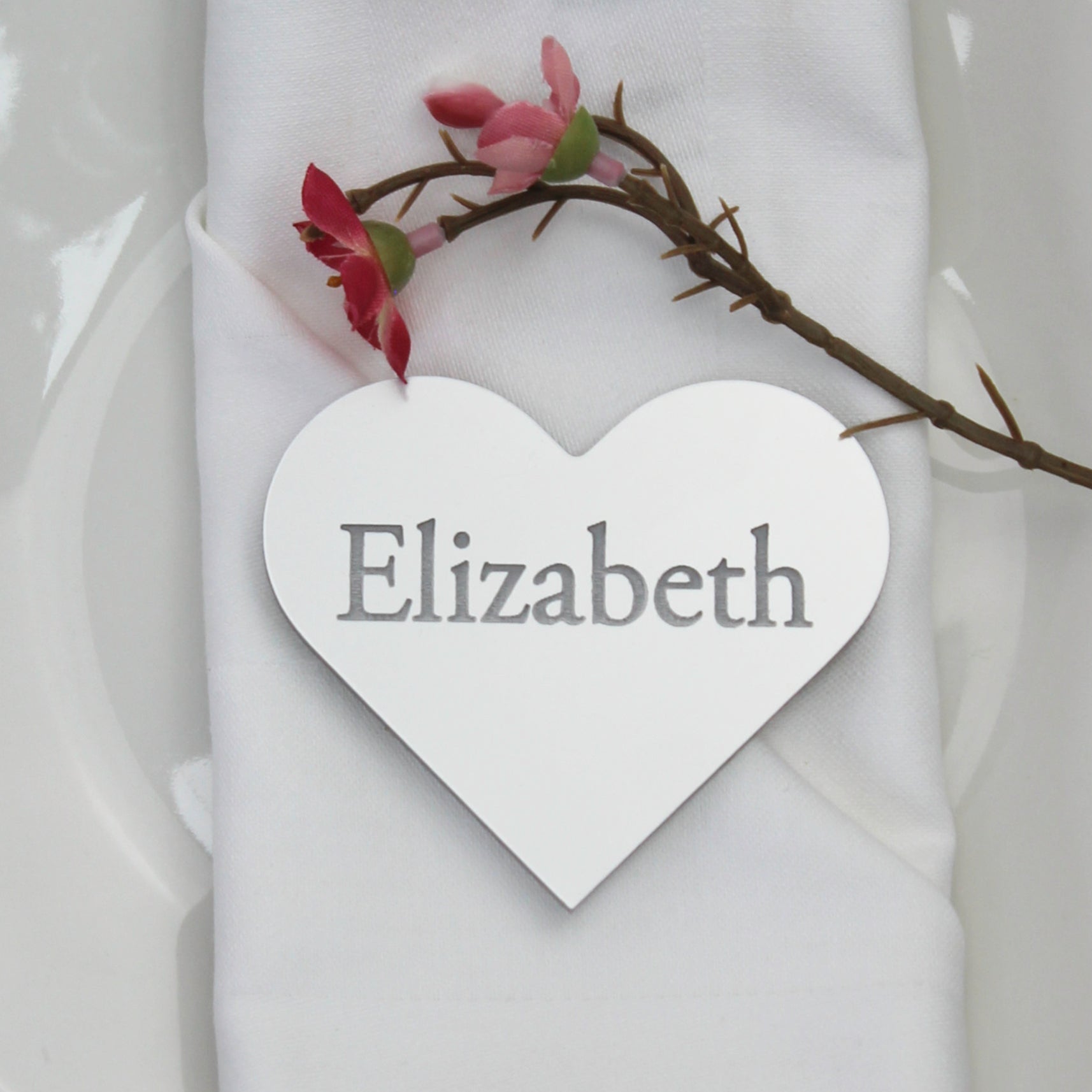 Heart-shaped place card with 'Elizabeth' on a folded white napkin with a branch and flowers on a gray tablecloth.