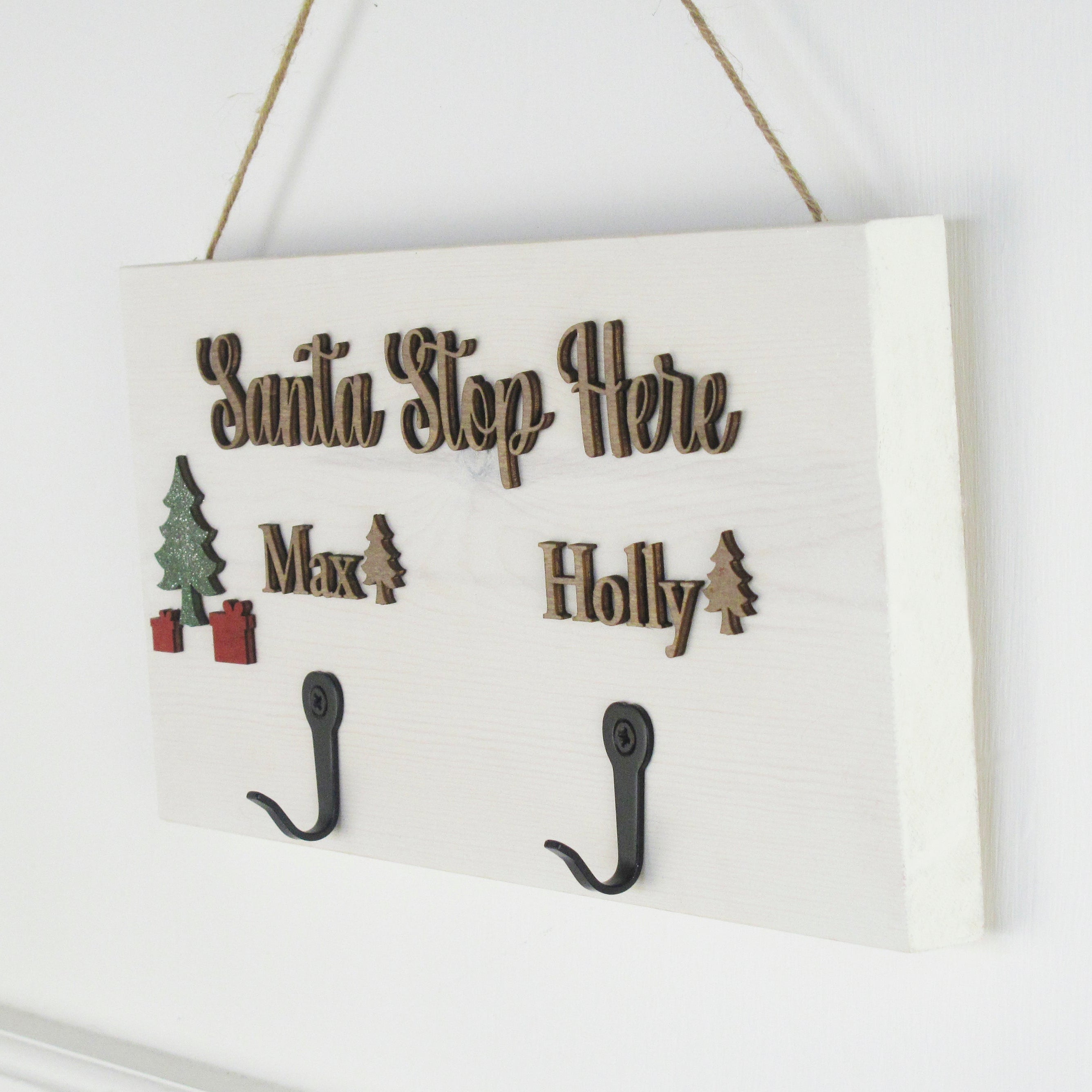 Decorative sign with 'Santa Stop Here' text and hooks for personalized names, hanging on a white wall.