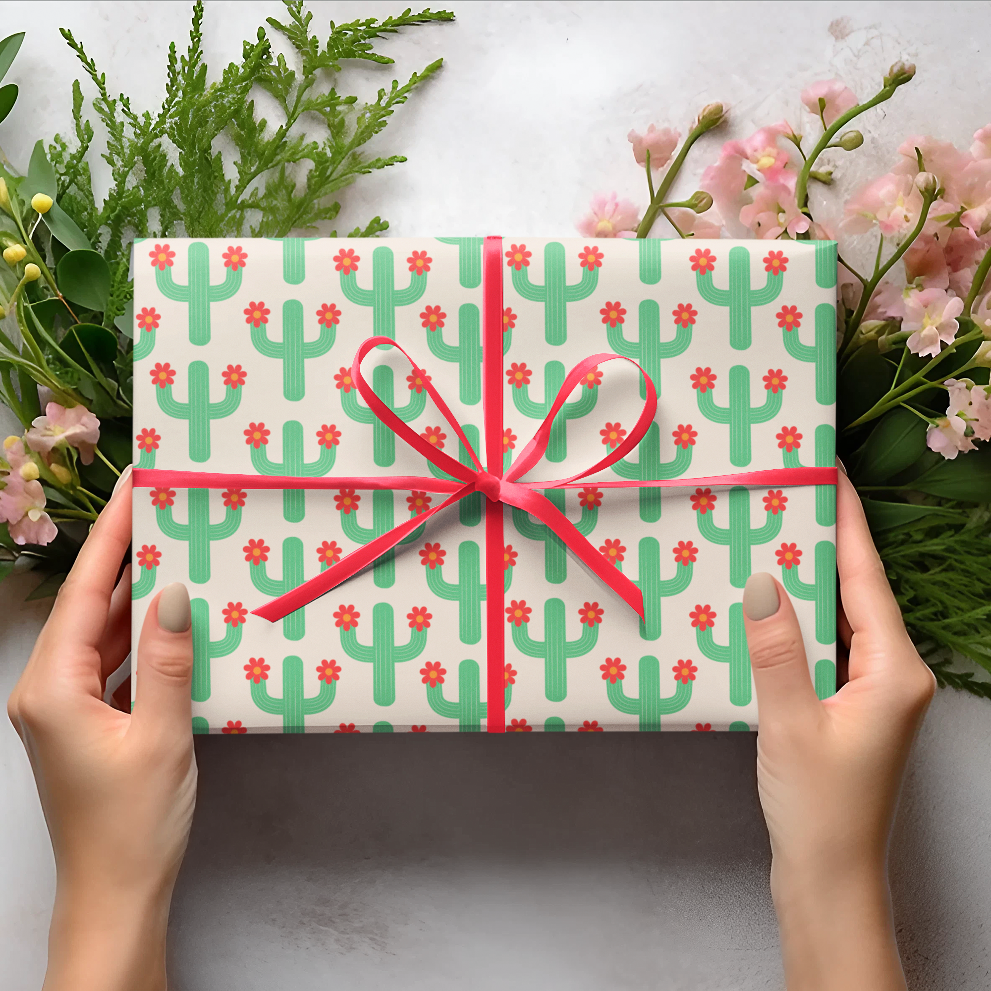 Gift box with green cactus pattern and red ribbon held by hands on a light background with flowers and leaves.