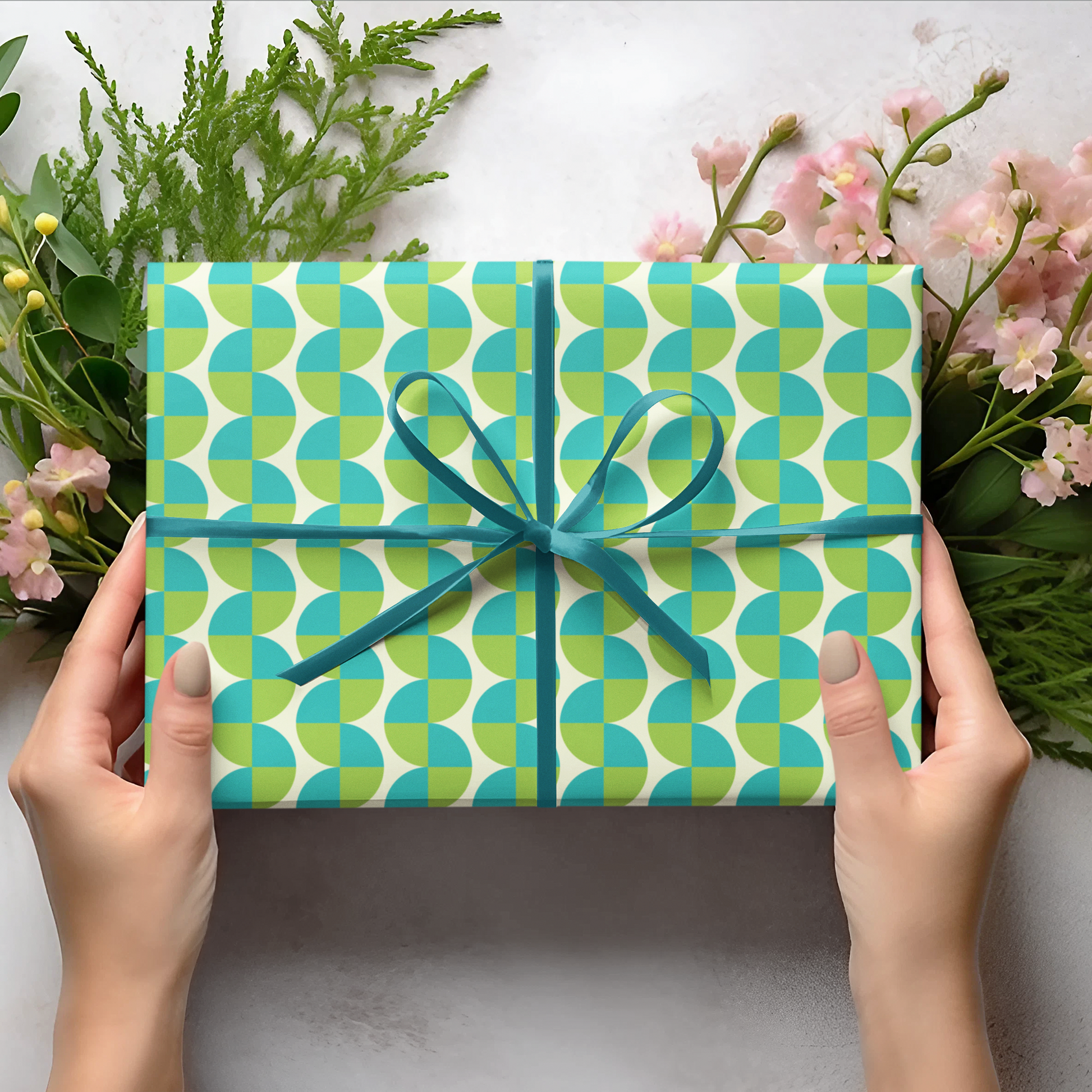 Gift box with green and blue pattern held by hands, surrounded by flowers and leaves on a light background