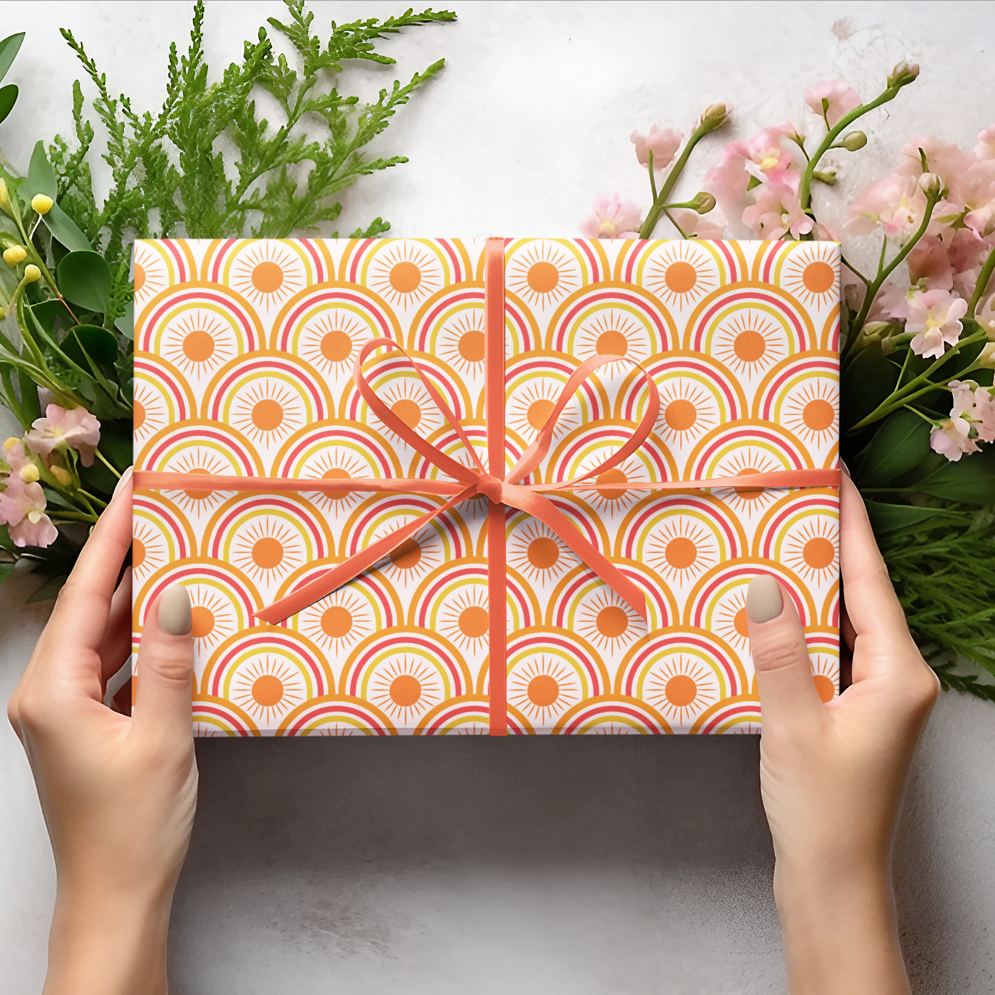 Gift box with orange geometric pattern held by hands, surrounded by greenery and flowers on a light background