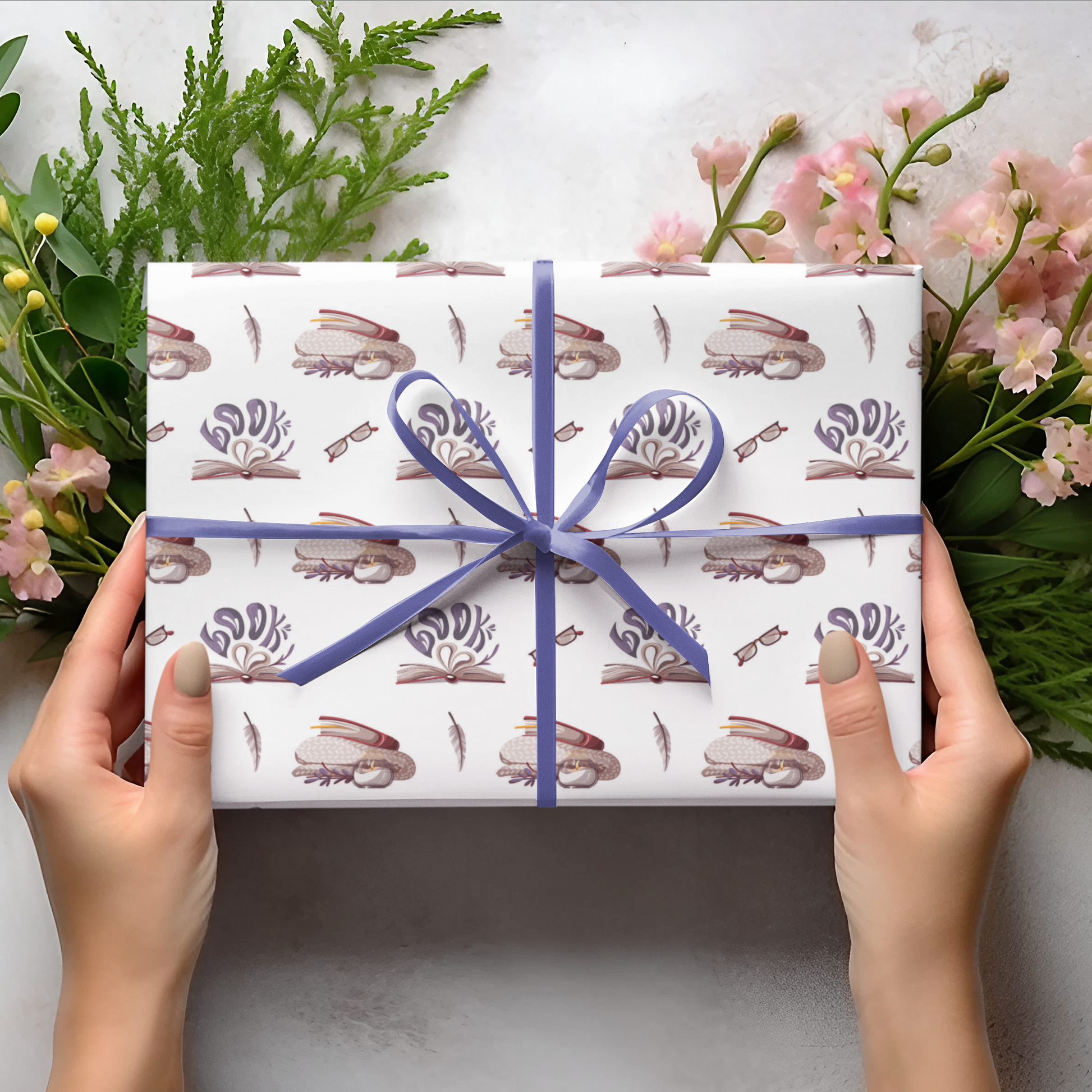Gift box with floral pattern and blue ribbon held by hands, surrounded by greenery and flowers on a light background