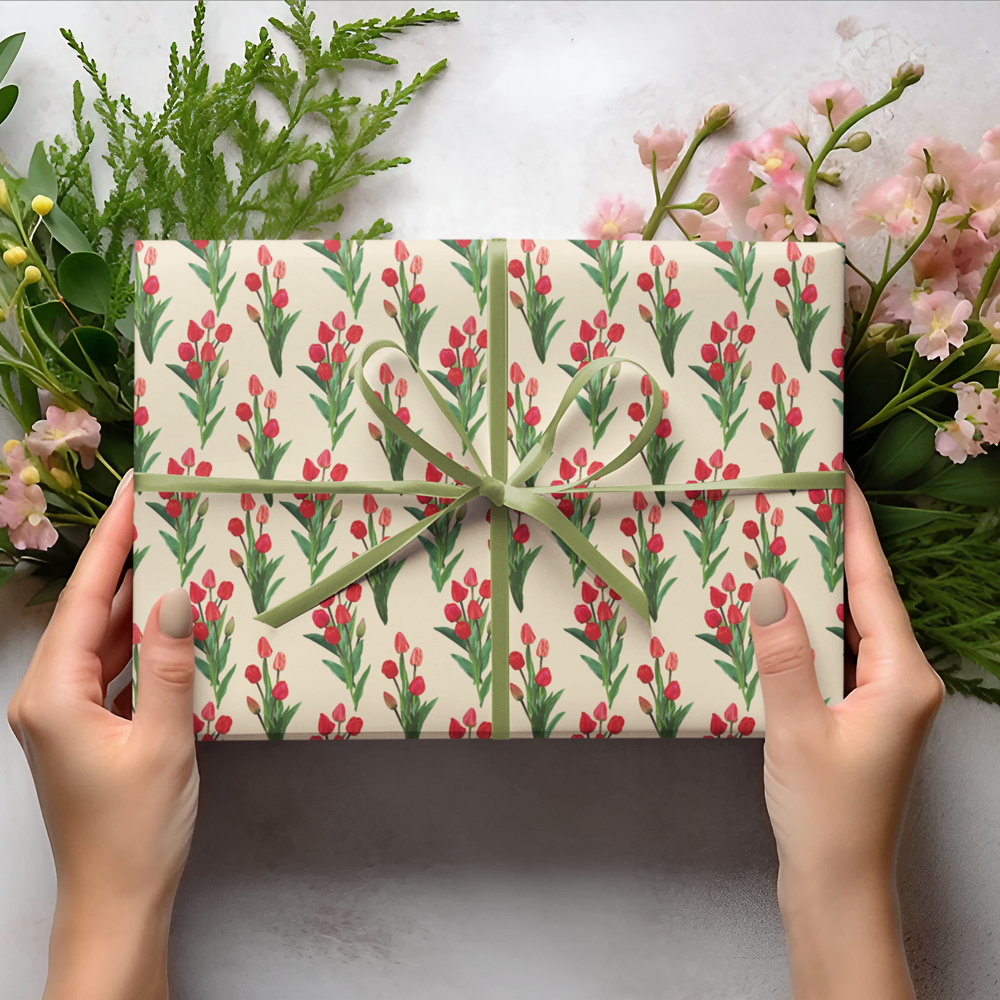 Gift box with floral pattern held by hands, surrounded by greenery and flowers on a light background