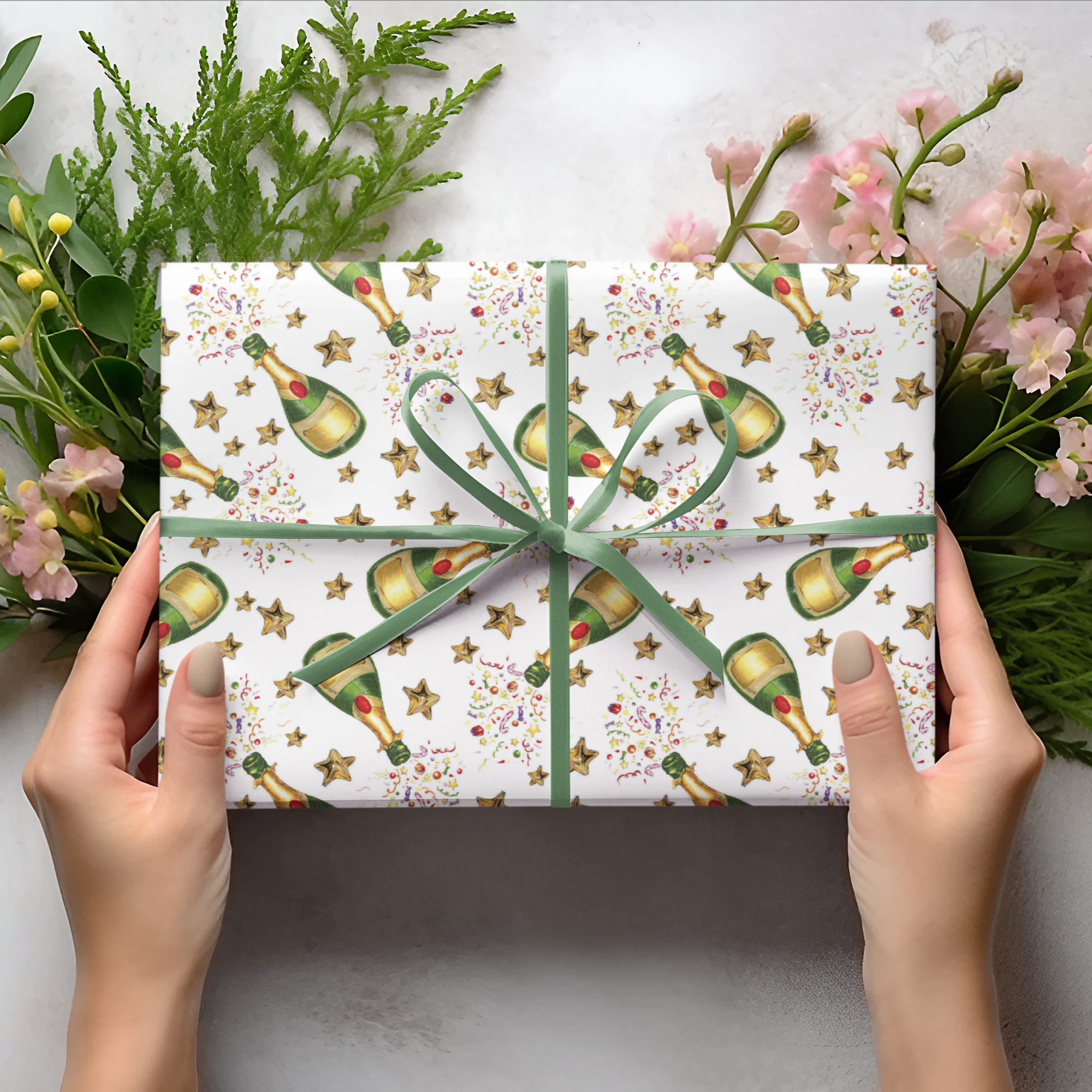 Gift wrap paper with champagne design held by hands, surrounded by flowers and greenery on a light background