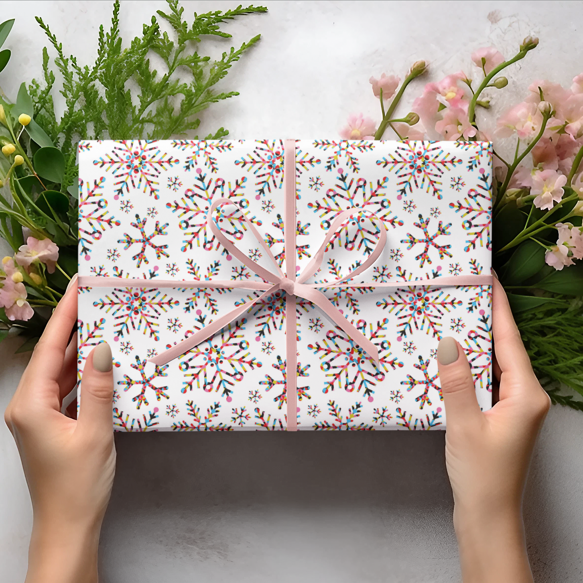 Gift wrap with snowflakes pattern held by hands on a light background with flowers and leaves.