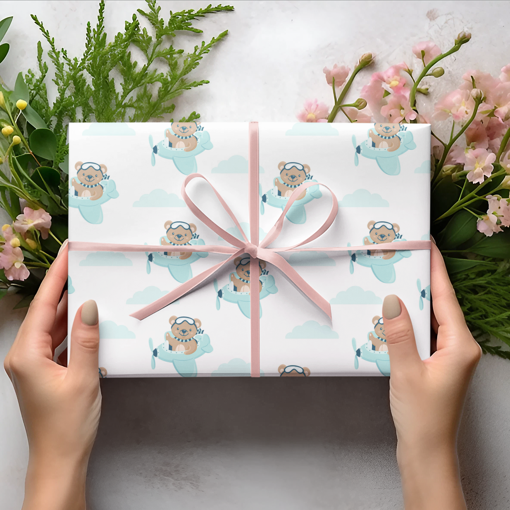Gift wrapping paper with teddy bear pattern held by hands, surrounded by flowers and greenery on a light background, perfect for little boys
