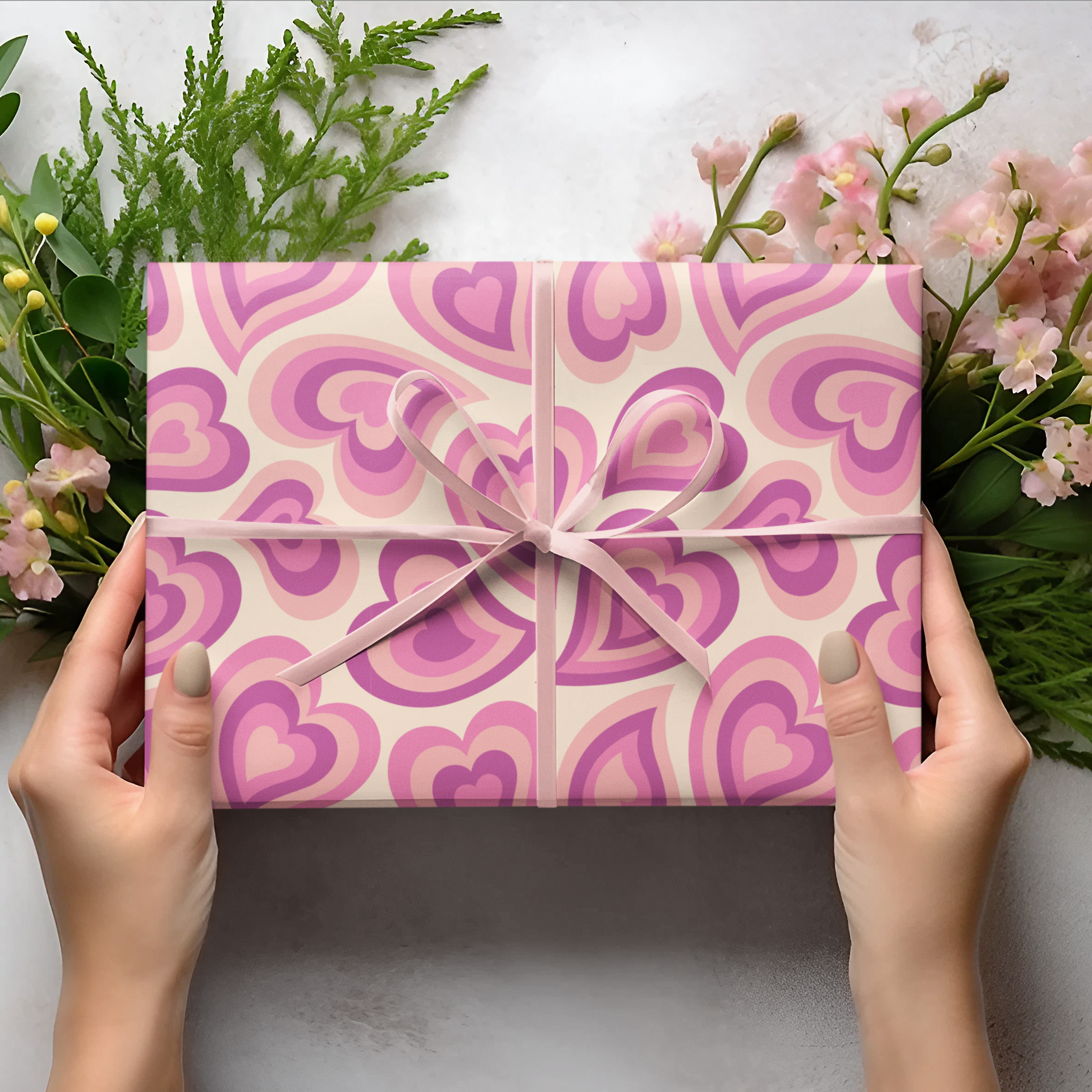 Pink gift box with heart pattern held by hands on a light background with flowers and leaves.