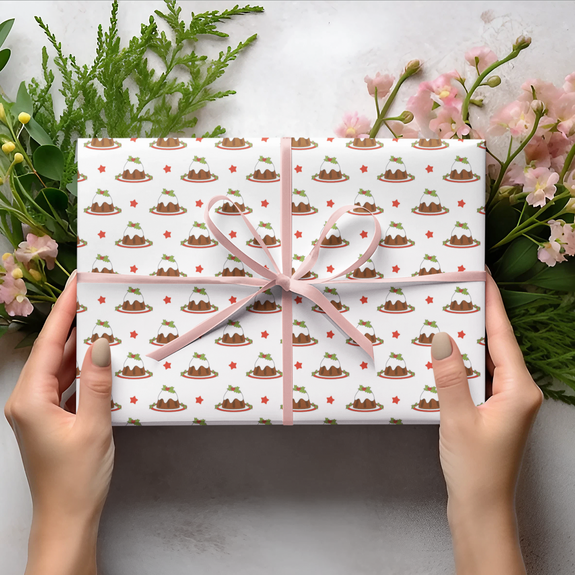 Christmas pudding gift wrap patterned paper and pink ribbon held by hands, surrounded by greenery and flowers on a light background